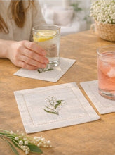 Load image into Gallery viewer, Bring quiet, botanical elegance to your table with these linen coasters embroidered with lily of the valley. Made from natural European linen, they protect surfaces beautifully while adding a refined spring-floral detail. Perfect for coffee moments, hosting, and thoughtful gifting.