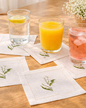 Load image into Gallery viewer, Bring quiet, botanical elegance to your table with these linen coasters embroidered with lily of the valley. Made from natural European linen, they protect surfaces beautifully while adding a refined spring-floral detail. Perfect for coffee moments, hosting, and thoughtful gifting.