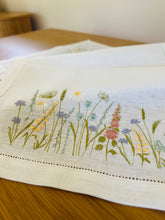 Ielādēt attēlu galerijas skatītājā, white linen table runner with machine‑embroidered meadow wildflowers motifs and hemstitched edges