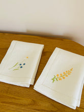 Ielādēt attēlu galerijas skatītājā, white linen napkins with machine‑embroidered meadow wildflowers motifs and hemstitched edges
